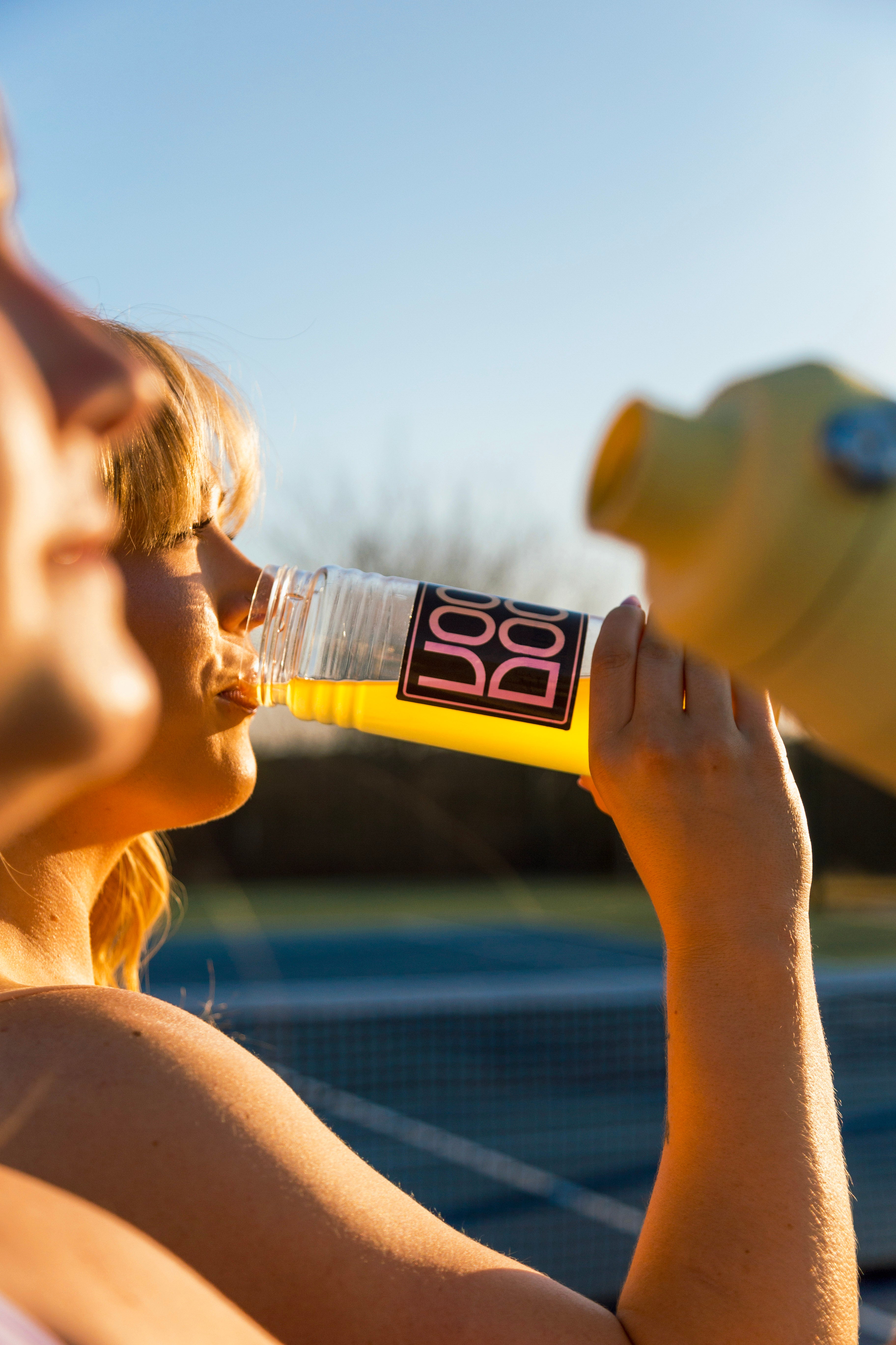 Person drinking yellow liquid out of a clear VooDoo water bottle of at a tennis court